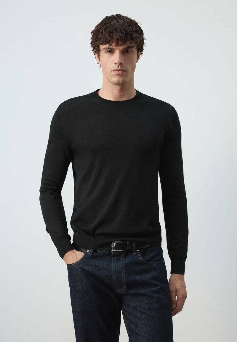 Homme aux cheveux courts et bouclés portant un pull noir à manches longues et un jean bleu foncé, debout avec une main dans la poche.