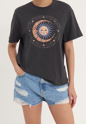 Person iført en sort t-shirt med sol- og månedesign og tekst, kombineret med revnede lyseblå denimshorts.