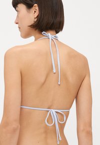 Šviesiai mėlynas halter stiliaus bikini viršus su plonomis dirželiais. Kaklo ir nugaros raišteliai, pasižymintis lygiu paviršiumi ir minimalistiniu dizainu, suteikiančiu klasikinį įvaizdį.