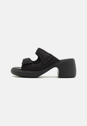 Black heeled mules with a soft fabric upper, padded straps, open toe, and wooden block heels, featuring a rounded shape and minimalist design.