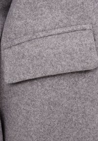 Šedá plsťová látka s texturovaným povrchem, vybavená obdélníkovou klopovou kapsou. Celkový design je jednoduchý a funkční.