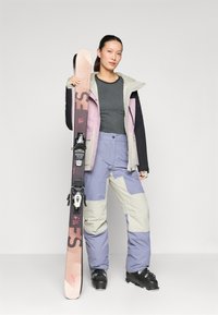 Skiausrüstung mit einem grauen Oberteil, einer mehrfarbigen Jacke in lila, schwarz und beige und helllila Hosen; hält Skier mit Bindungen.