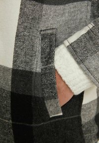 Szaro-czarny flanelowy płaszcz w kratę z białym polarowym podszyciem. Posiada boczną kieszeń i miękką fakturę, podkreślającą ciepło i wygodę.