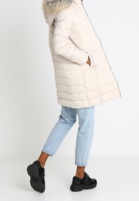 Beige puffermantel med huva, prydd med fuskpäls, quiltat mönster, sidofickor och något längre bak. Bärs med ljusblå jeans och svarta sneakers.