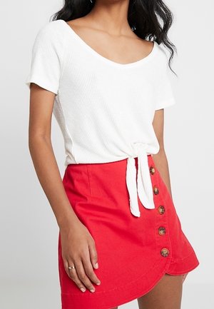 Biała prążkowana bluzka z krótkim rękawem i wiązaniem z przodu, w zestawie z czerwoną spódnicą kopertową z brązowymi guzikami po boku.