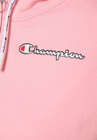 Ljusrosa sweatshirt med dragkedja i halsen, med vit dragsko och broderad svart "Champion"-logotyp med röd accent. Mjuk textur.
