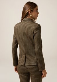 Blazer marron à motif avec une coupe ajustée, des épaules structurées et une fente dans le dos. Le tissu présente un imprimé texturé et complexe.
