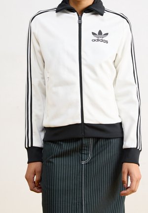 Personne portant une veste zippée blanche Adidas avec des rayures noires sur les manches et un col noir, associée à une jupe sombre à fines rayures.