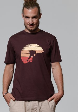 Homme aux cheveux attachés portant un T-shirt bordeaux avec la silhouette d'un grimpeur escaladant une falaise sur fond de coucher de soleil.