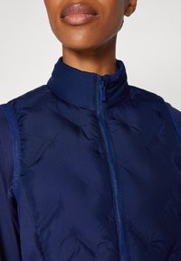 Tamno plavi puffer prsluk s visokim ovratnikom, koji ima puni patentni zatvarač, teksturiranu tkaninu i glatke, podstavljene sekcije za izolaciju.