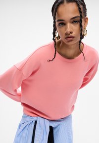 Rosa hettegenser med en avslappet passform og senkede skuldre, med en teksturert stoff. Kombinert med en blåstripete skjorte knyttet i livet.