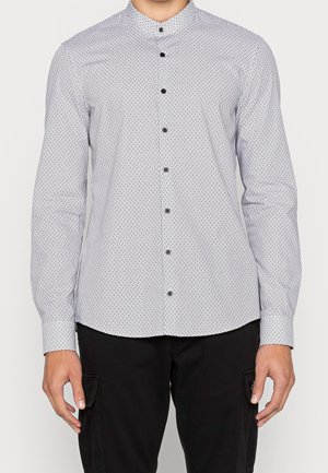 Person trägt ein eng anliegendes weißes Langarmshirt mit kleinen schwarzen geometrischen Mustern und schwarze Hosen mit Seitentaschen.