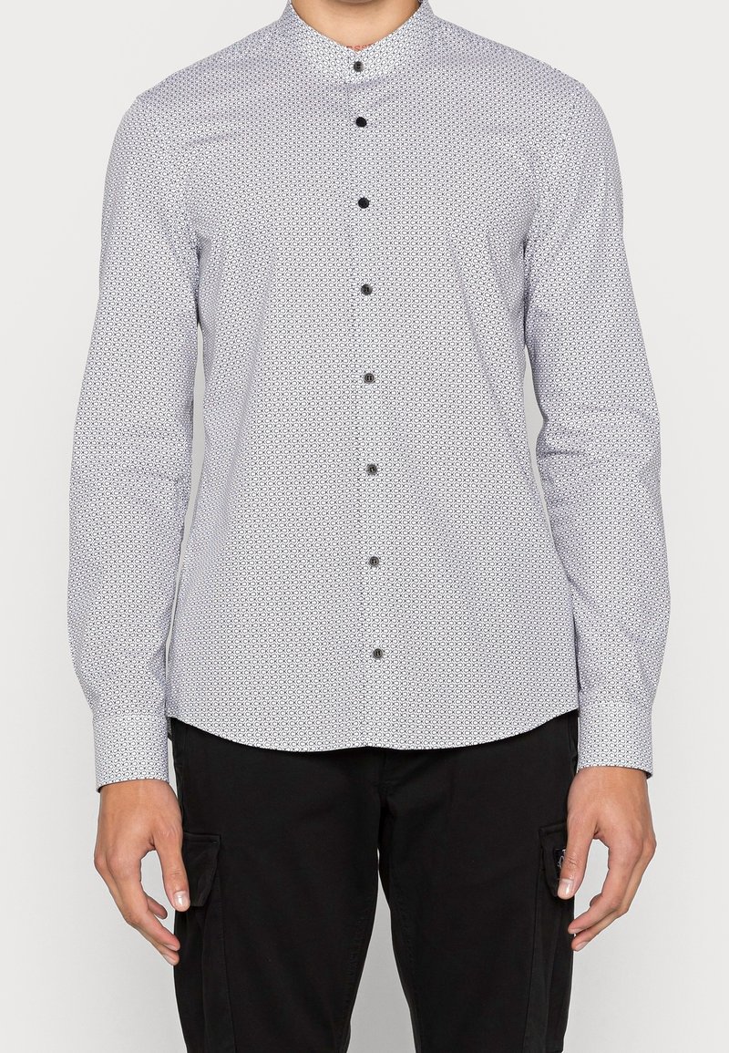 Personne portant une chemise blanche cintrée à manches longues avec de petits motifs géométriques noirs et un pantalon noir avec des poches latérales.