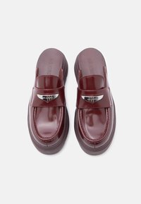 Μπορντό παντόφλες από γυαλιστερό πατεντάκι, με επίπεδη πλατφόρμα και μεταλλικές λεπτομέρειες στη λουρίδα.