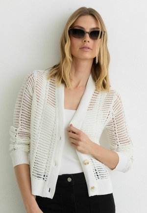 Blonde Frau mit dunkler Sonnenbrille, weißem Netztop über weißem Oberteil und schwarzer Hose, posiert vor einer schlichten hellen Wand.