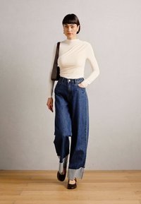 Maglia bianca a maniche lunghe con design asimmetrico, abbinata a pantaloni in denim blu a vita alta e gamba larga con polsini a contrasto e scarpe nere.