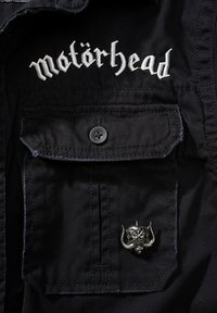 Juodas medvilnės švarkas su sidabro spalvos "Motörhead" siuvinėjimu ant apykaklės, turintis mygtukinę kišenę ir metalinį kaukolės smeigtuką.