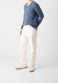 Maglione lavorato a maglia blu a scollo a V, pantaloni bianchi dalla vestibilità comoda e sneakers bianche con suole marroni chiari. Il maglione presenta un logo discreto sul petto.