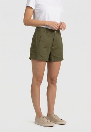 Femme portant un short taille haute vert olive avec des poches latérales, un t-shirt blanc à manches courtes et des baskets beige sur un fond uni.
