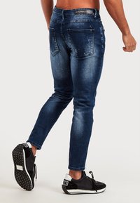 Slim-fit mörkblå denimjeans med en blekt yta och subtila slitningar. Fem fickor med ett märkesbälte. Bärs med svarta sneakers.