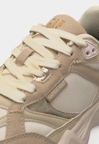 Gros plan sur une basket beige et crème avec des lacets crème, en suède et mesh, et une étiquette "GAP EST.1969" près des lacets.
