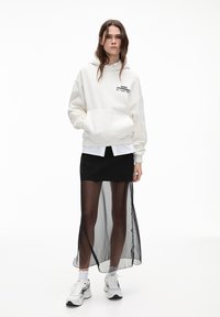 Sweatshirt blanc surdimensionné avec poche, jupe noire avec superposition transparente, associée à des baskets blanches et superposée avec une chemise noire et blanche.