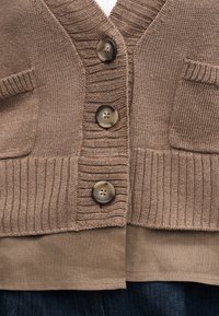 Καφέ πλεκτή ζακέτα με τρία κουμπιά, δύο μικρές μπροστινές τσέπες, ριμπ γιακά και τελείωμα, φορεμένη πάνω από μία μπεζ πουκάμισο και σκούρο παντελόνι.