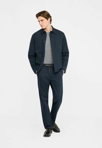 Chemise bleu marine boutonnée, portée sur un pull gris, associée à un pantalon bleu marine assorti et des chaussures noires. Tissu lisse, coupe décontractée, design minimaliste.
