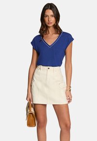 Blauw V-hals T-shirt met korte mouwen, gecombineerd met een crèmekleurige mini-jurk met voorzakken en riemlussen. Model houdt een bruine handtas vast.