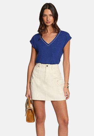 Chemise bleue à col V avec des manches courtes, associée à une mini-jupe crème dotée de poches avant et de passants de ceinture. Le modèle tient un sac à main marron.