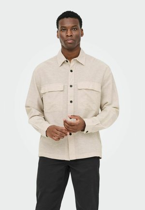 Homme portant une chemise beige à boutons avec poches à rabat et pantalon noir, debout devant un fond gris clair.