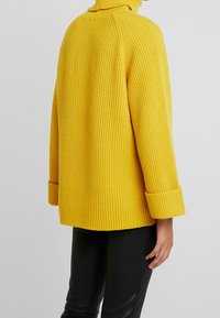Žlutý žebrovaný svetr s vysokým límcem a ohrnutými manžetami. Vyrobený z texturovaného materiálu, končící v délce boků s volným střihem.