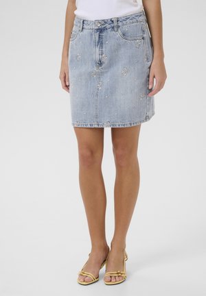 Femme portant une jupe en jean brodée bleu clair, un haut blanc et des talons ouverts beige, debout devant un fond blanc uni.