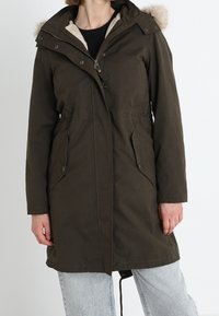 Parka de color verde oliva con capucha forrada de piel, cierre frontal de cremallera y dos bolsillos laterales. Cuenta con cintura y dobladillo ajustables.