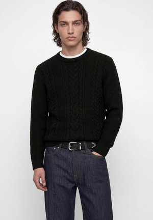 Un mannequin masculin se tient devant un fond blanc, vêtu d’un pull noir en maille torsadée, d’un jean foncé et d’une ceinture noire.