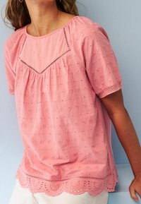 Blouse rose clair à manches courtes, avec un motif floral brodé, un ourlet festonné et des détails en dentelle autour du col et des manches.