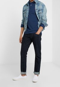 Man som bär en ljusblå denimjacka över en marinblå pikétröja, mörka uppvikta jeans och vita slip-on sneakers, står med en hand i fickan.