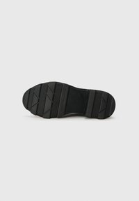 Semelle de chaussure en caoutchouc noir avec une surface texturée et des rainures horizontales et diagonales marquées, arborant un logo de marque sur le côté.