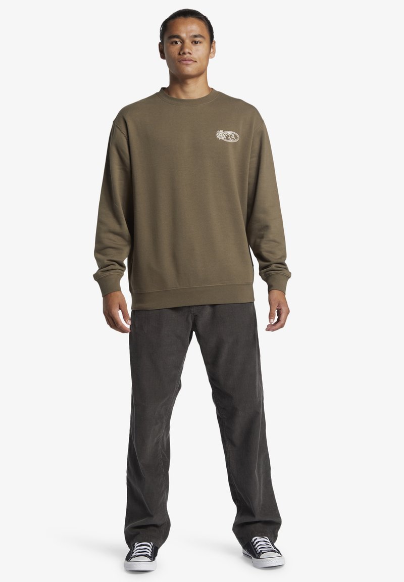 Olijfgroene sweatshirt met ronde hals en lange mouwen; heeft een klein wit logo op de borst. Gecombineerd met donkergrijze corduroy broek en sneakers.
