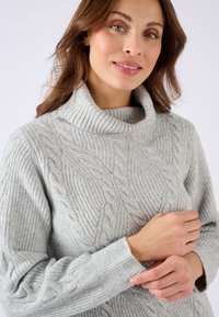 Suéter de cuello alto gris claro hecho de un tejido texturizado, que presenta un diseño de punto de cables con puños y cuello acanalados.