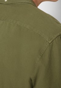 Gros plan du haut du dos d'une personne portant une chemise en tissu texturé vert olive avec des coutures visibles.