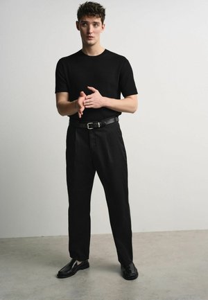 Noor mees seisab toas, kandes musta lühikeste varrukatega soonikulehte, musta vööd, musti pükse ja musta poleeritud jalatseid.