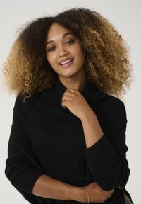 Maglione nero a maglia con una texture a coste e colletto ampio, indossato da una persona con capelli ricci e colpi di luce. Gioielli minimalisti accentuano il look.