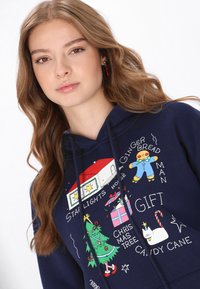 Tengerészkék kapucnis pulóver ünnepi grafikával, amelyen mézeskalács figura, karácsonyfa, ajándékok és élénk színű ünnepi motívumok láthatók.
