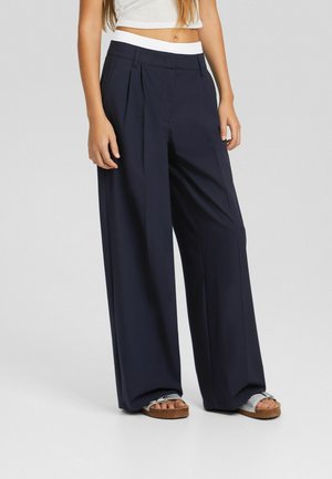 Femme portant un pantalon large taille haute de couleur bleu marine foncé avec des plis sur le devant et une ceinture blanche, assorti à des sandales argentées et un haut blanc court.