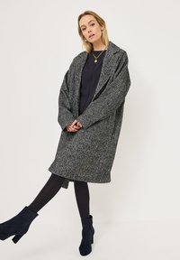 Nadměrný kabát v šedém houndstooth vzoru s klopami, strukturovaná tkanina a boční kapsy. V kombinaci s černými punčochami a námořnickými kotníkovými botami.