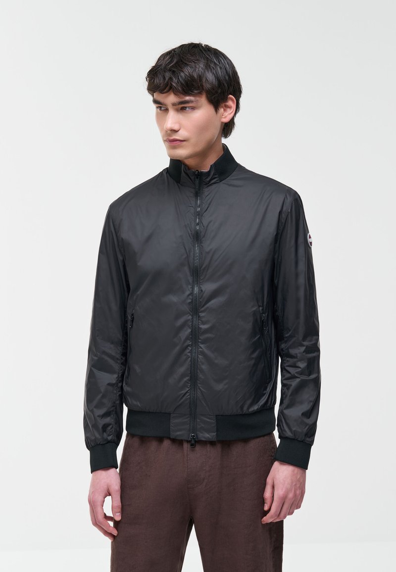 Junger Mann trägt eine schwarze Zip-Up-Jacke mit gerippten Bündchen und Kragen, stehend vor einem einfarbigen hellen Hintergrund.