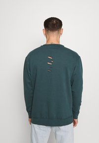 Tyrkysová mikina s dlouhými rukávy, kulatým výstřihem a pěti vertikálními trhlinami na zadní straně. Má volný střih a žebrované detaily na manžetách a lemu.