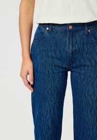 Jeans di denim blu con motivi a spirale in rilievo, caratterizzati da un classico design a cinque tasche e dettagli in rame nella vita.