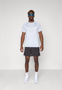 Bărbat îmbrăcat în echipament sportiv, cu tricou alb, pantaloni scurți negri, adidași albi, șosete și ochelari de soare sportivi albaștri, stând pe un fundal simplu.